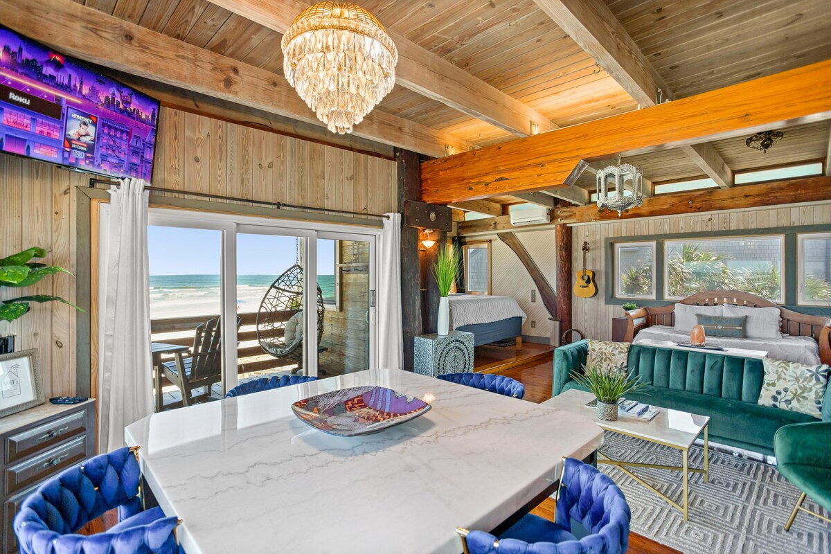 An open living area features wooden beams and natural light streaming through large glass doors leading to a beachfront balcony. A modern dining table is surrounded by blue velvet chairs, while a plush teal sofa and a chandelier accentuate the artistic interior.
