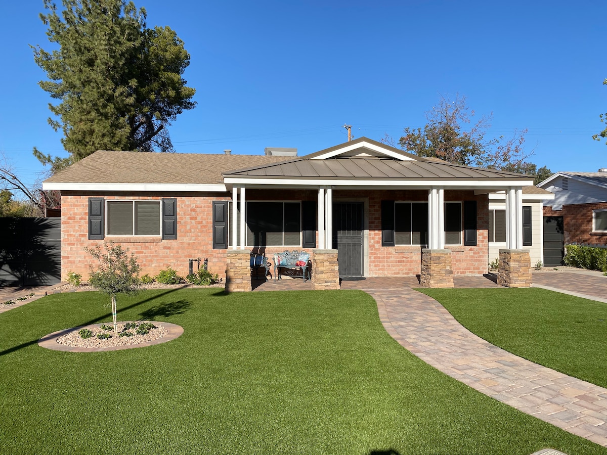 The charming exterior features a brick façade with a covered front porch supported by classic columns. A manicured lawn and stone pathway lead to the entrance. Dark window shutters add contrast to the sunny atmosphere, while surrounding trees provide a touch of greenery.