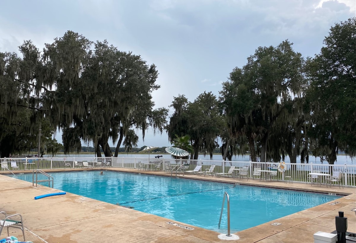 A clear swimming pool is bordered by a spacious deck with lounge chairs under shaded trees. Spanish moss hangs from the oaks, providing a natural backdrop. The tranquil lake can be seen in the distance, adding to the serene environment.