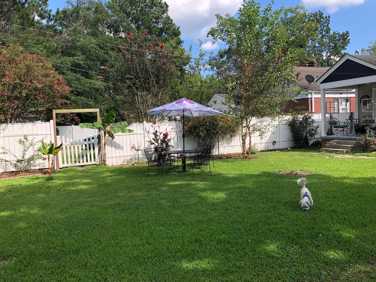A spacious backyard is framed by a white fence, featuring a patio set with a colorful umbrella. Lush green grass surrounds the area, complemented by blooming flowers and shrubs. A small dog explores the yard, adding a lively element to the serene outdoor space.