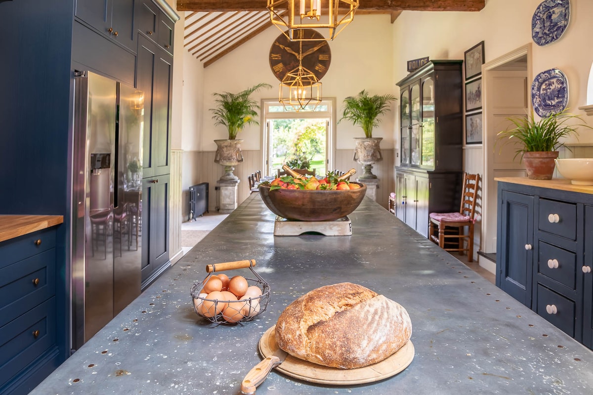 A spacious kitchen features a large central island topped with a loaf of bread and a wooden board. A bowl filled with fresh fruit is centered on the island, while stainless steel appliances complement the rich blue cabinetry. Natural light floods in through a large window.