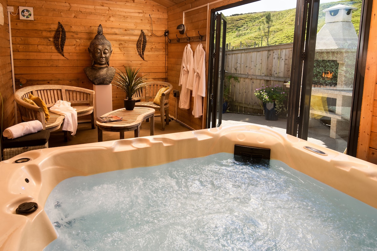 An inviting spa area features a luxurious hot tub surrounded by wooden paneling. Natural light floods the space, with large glass doors opening to the outdoor area. A relaxed seating arrangement includes two wicker chairs and a small table, enhancing the serene atmosphere.