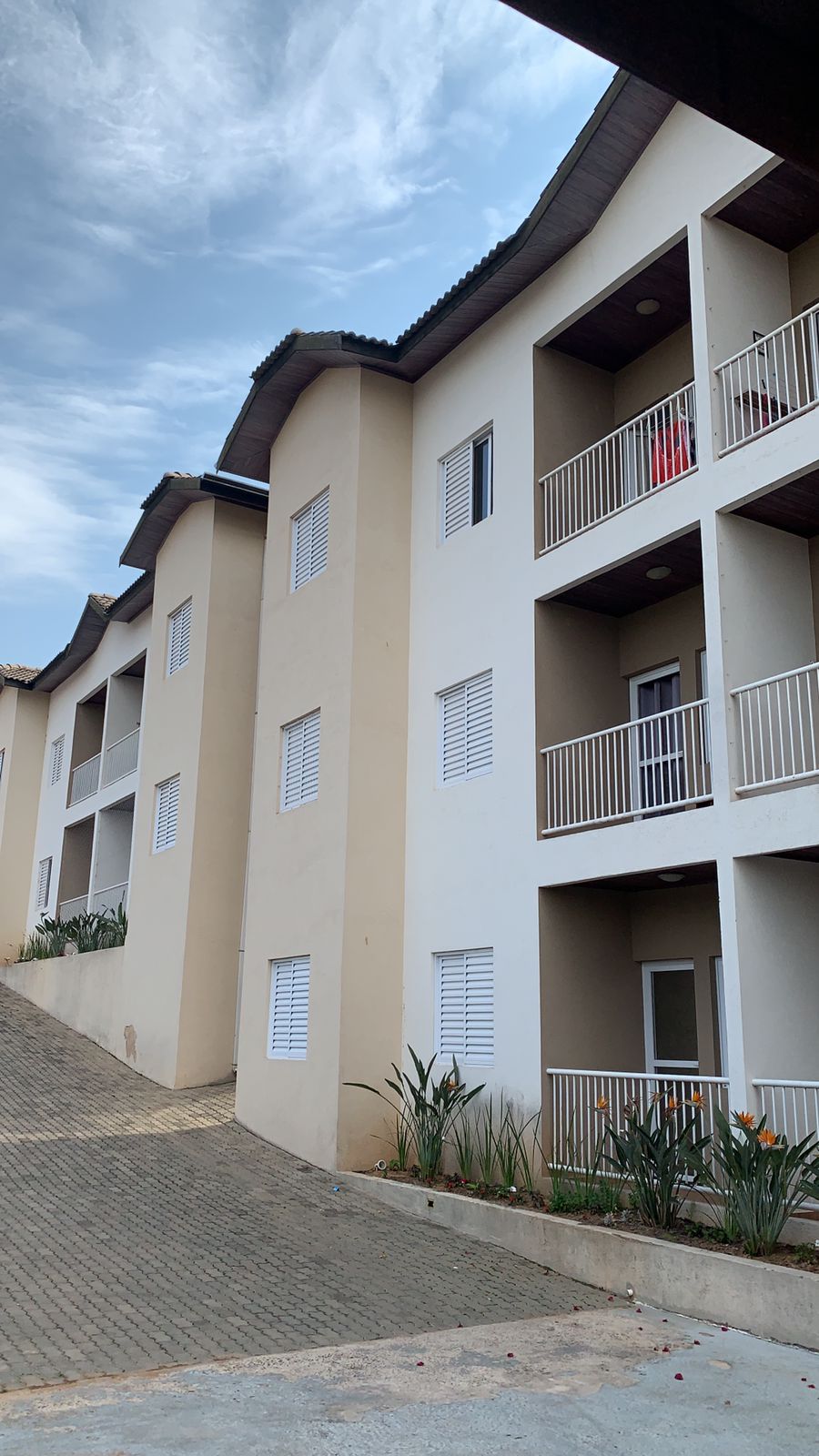 The image shows a multi-story building with a modern architectural design, featuring balconies and white exterior walls. Paved pathways lead up to the entrance, with plants lining the base of the structure. The cloudy sky offers a soft, diffused light over the scene.