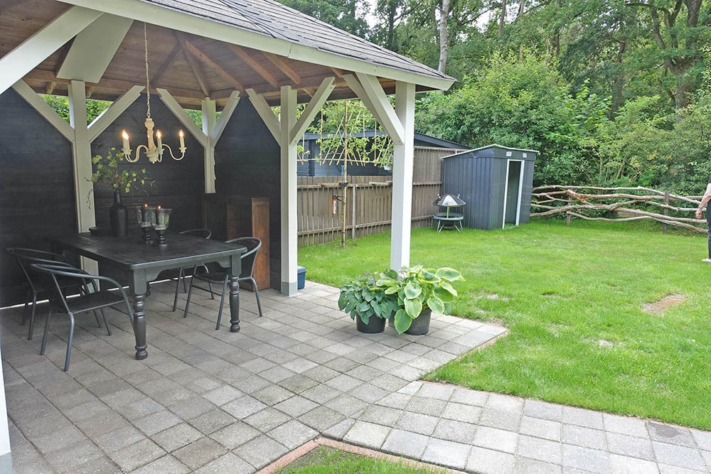 A shaded outdoor patio features a wooden table surrounded by chairs. A chandelier is suspended above the table, adding elegance to the space. Lush greenery and neatly arranged hedges frame the area, with a well-maintained lawn visible in the background.