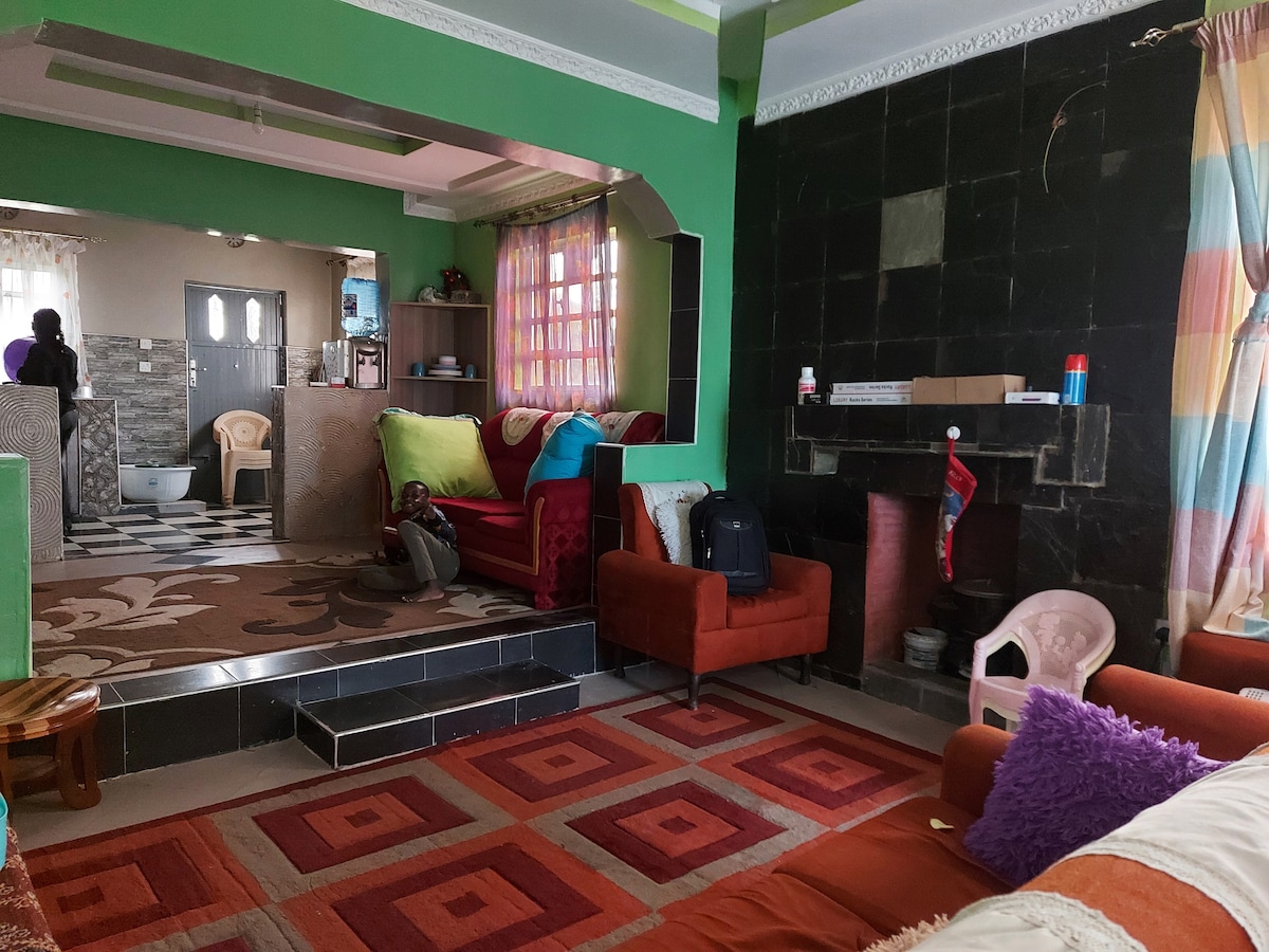 The living area features a combination of vibrant green and orange walls, with comfortable seating arranged around a fireplace. A patterned rug enhances the cozy atmosphere. A doorway leads to a bathroom visible in the background, while colorful curtains add brightness to the space.