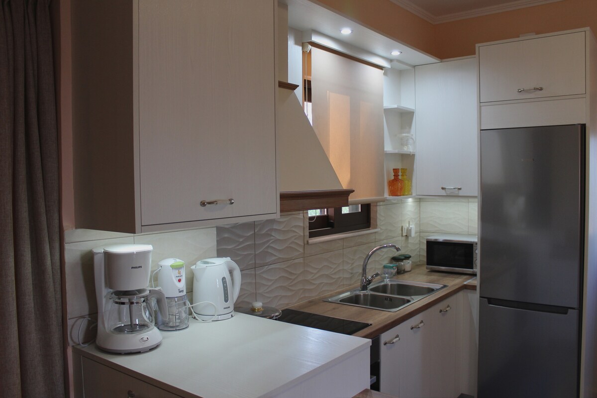 A well-equipped kitchen is visible, featuring white cabinetry, a stainless steel refrigerator, and a built-in microwave. Countertops are clean and organized, with a coffee maker and kettle on display. Natural light filters through a window, enhancing the functional layout.