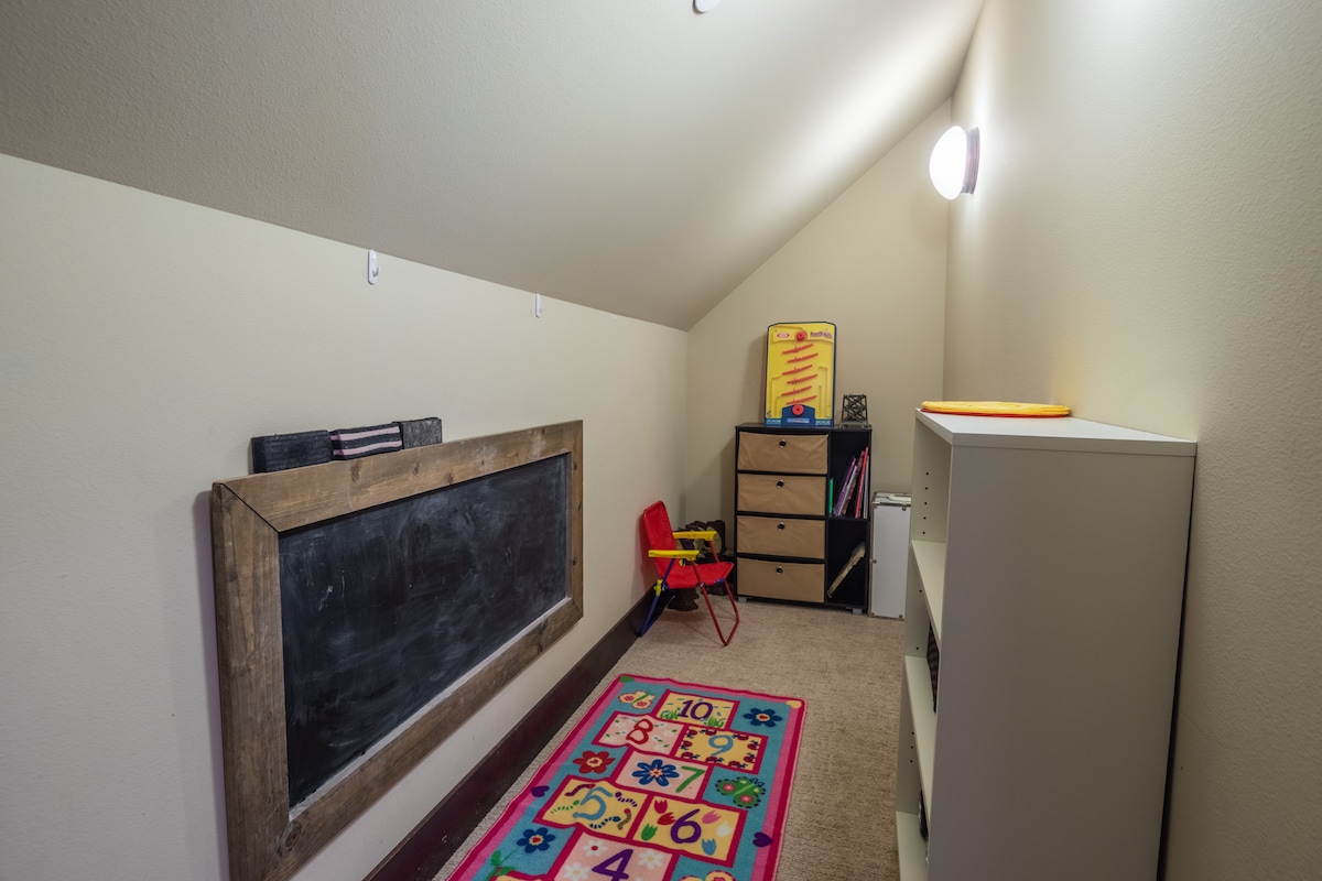 An attic playroom is equipped with a chalkboard and a brightly colored area rug with numbers. A small red chair is positioned near a storage unit containing various items. Soft lighting enhances the cozy atmosphere of this child-friendly space.