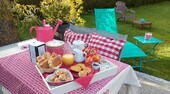 A dining table is set outdoors, featuring a variety of baked goods, fruits, and beverages in aesthetically arranged containers. The scene includes patterned textiles and colorful seating options, surrounded by greenery, creating a relaxed setting for enjoying breakfast.