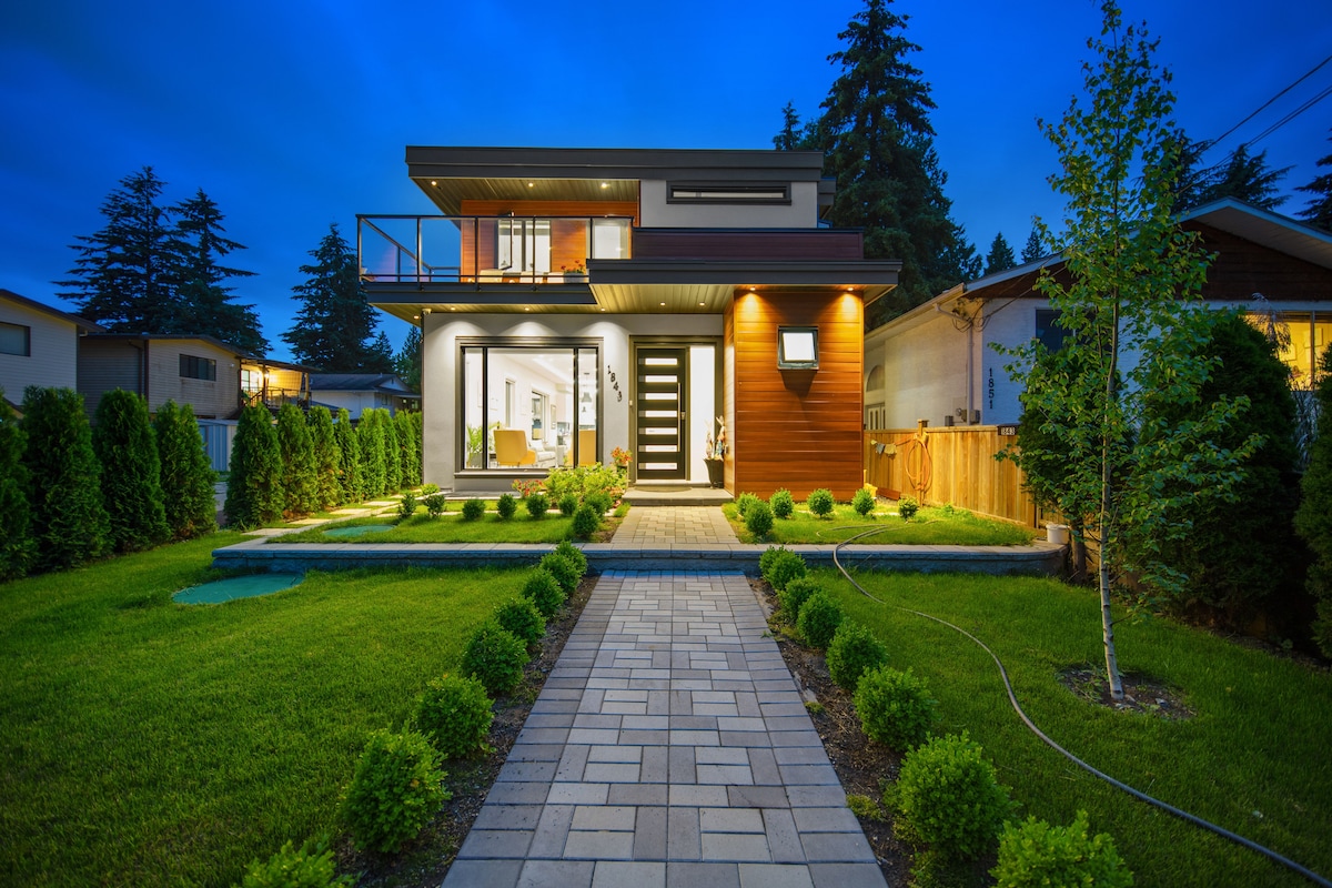 The exterior of a modern guest suite is illuminated in the evening light. A pathway of pavers leads to a welcoming entrance, bordered by neatly trimmed hedges. Large glass doors create a connection to the outdoor space, enhancing the inviting facade of the home.