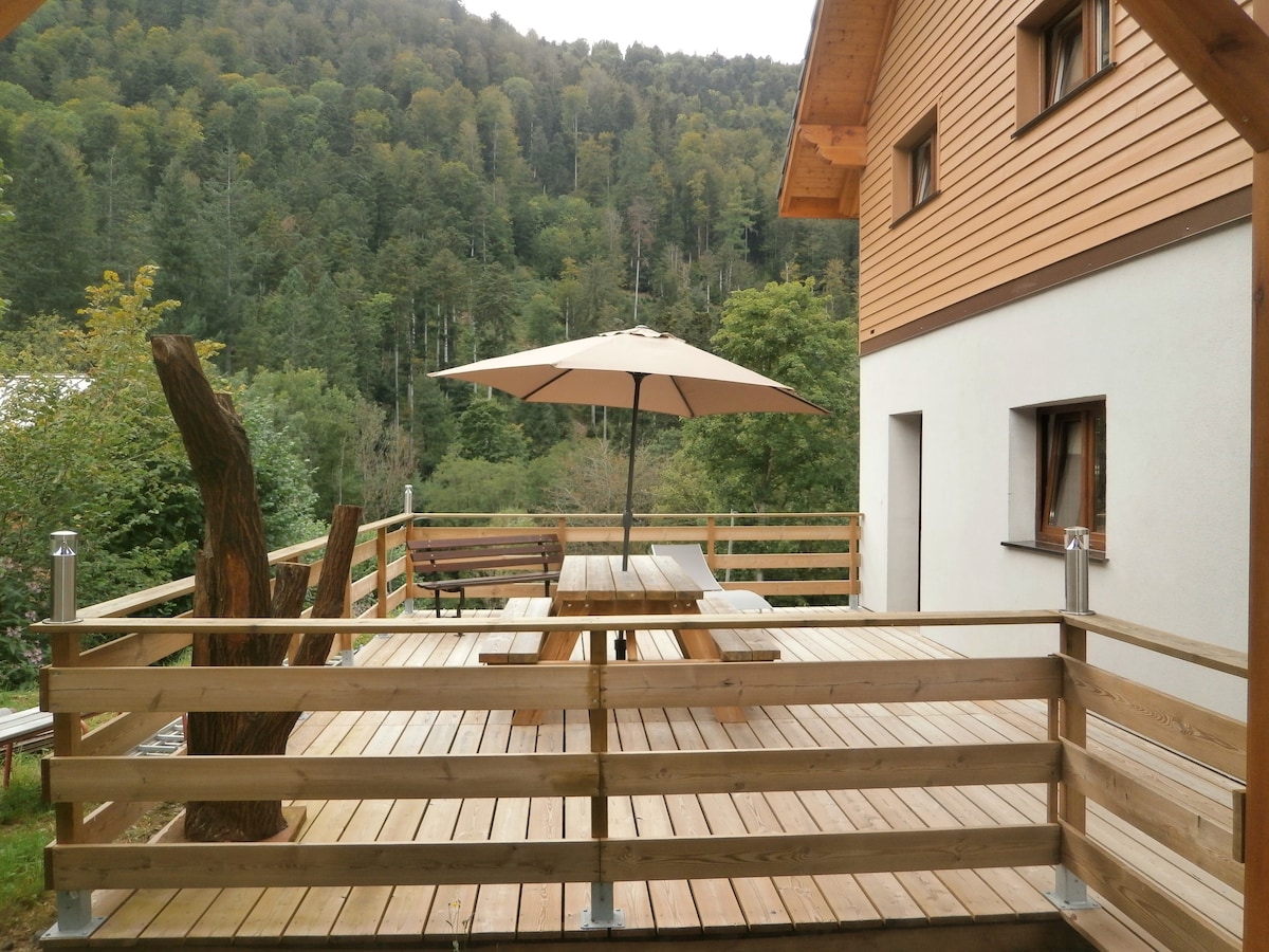 A wooden deck area is featured, furnished with a large picnic table under a beige umbrella. A bench is positioned nearby, and the space is surrounded by a wooden railing. Lush greenery of trees is visible in the background, complementing the natural setting.
