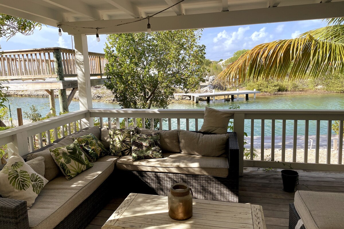 A covered outdoor terrace features a comfortable seating area with plush cushions and a rustic wooden table. Lush tropical foliage frames the scene, with water visible in the background, creating a serene setting for relaxation. Soft lighting is suspended above, enhancing the tranquil ambiance.