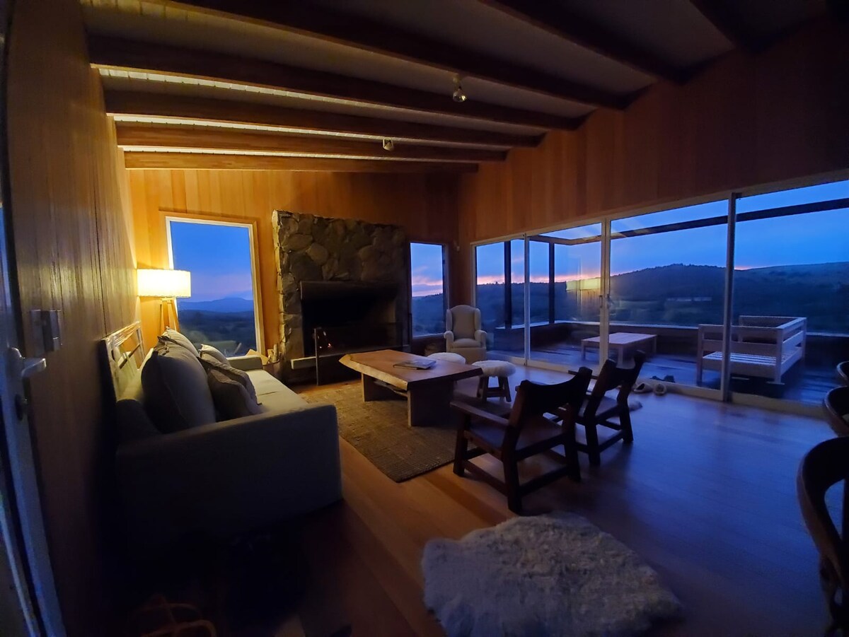 A modern living area features a wooden wall and large windows showcasing scenic views at dusk. A stone fireplace complements a light-colored sofa and a wooden coffee table. Soft lighting is provided by a floor lamp, and wooden chairs are arranged nearby.