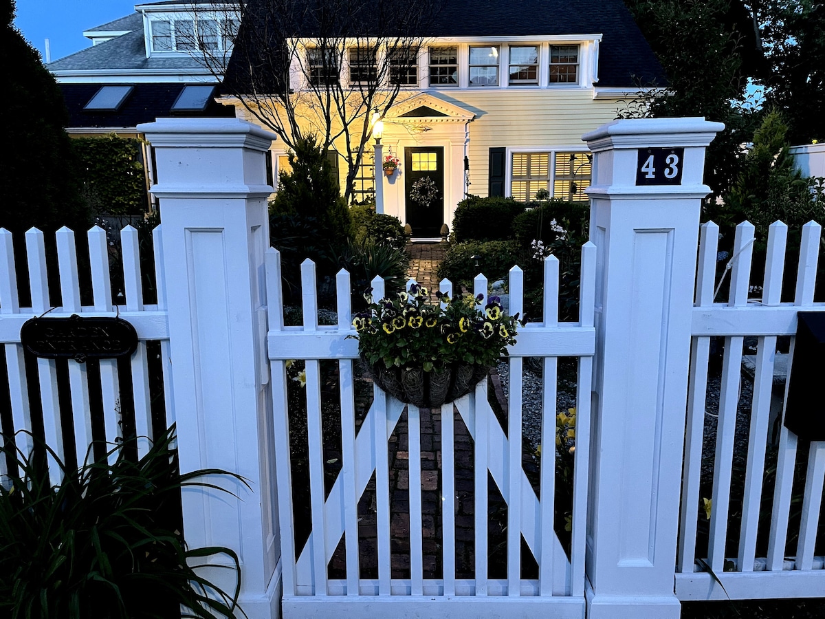 A white picket fence frames the entrance, featuring a decorative planter with flowers. The welcoming front door is partially visible, situated beneath a classic porch light. Surrounding greenery adds a touch of nature to the charming exterior, gently illuminated in the evening light.