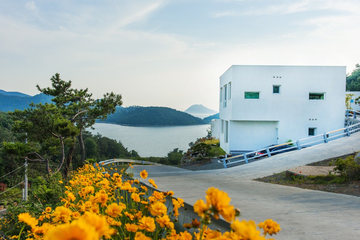 A modern white structure is positioned on a hillside overlooking a serene body of water, framed by lush greenery and distant mountains. Bright yellow flowers line the pathway leading to the entrance, enhancing the natural beauty of the surroundings.