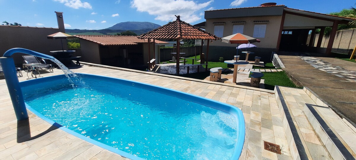 A sparkling blue pool features a gentle waterfall, surrounded by tiled patio and grassy areas. Nearby, outdoor seating with umbrellas offers shaded spots for relaxation. In the background, an open-air gazebo and a spacious building can be seen against a scenic mountain backdrop.
