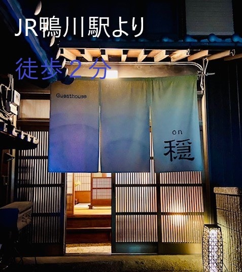 The entrance of the guesthouse is highlighted by a pair of fabric curtains, displaying the name 'on穏'. Traditional wooden sliding doors and a stone pathway are visible, creating a welcoming entrance just a short walk from the nearby train station.