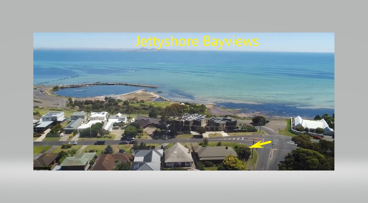 Aerial view showcases the bay with its calm blue waters and sandy beach. Surrounding properties and lush greenery line the shoreline, while the road leads towards the beach, indicating accessible pathways for visitors. The tranquil scene offers an expansive outlook over the coastline.