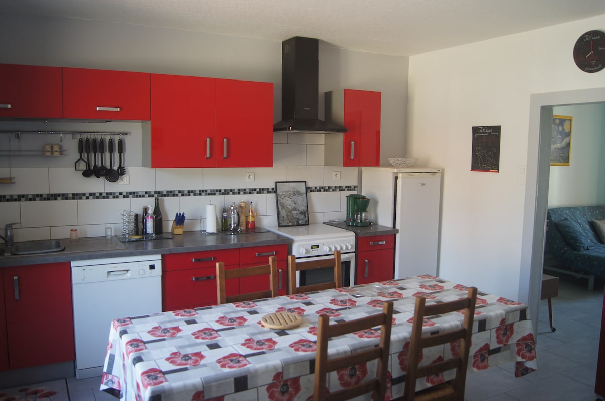 A modern kitchen features bright red cabinets and a spacious dining table with a floral tablecloth. Appliances include a stove, refrigerator, and dishwasher. Cookware and utensils are neatly arranged, while a seating area is visible in the background with a green couch.