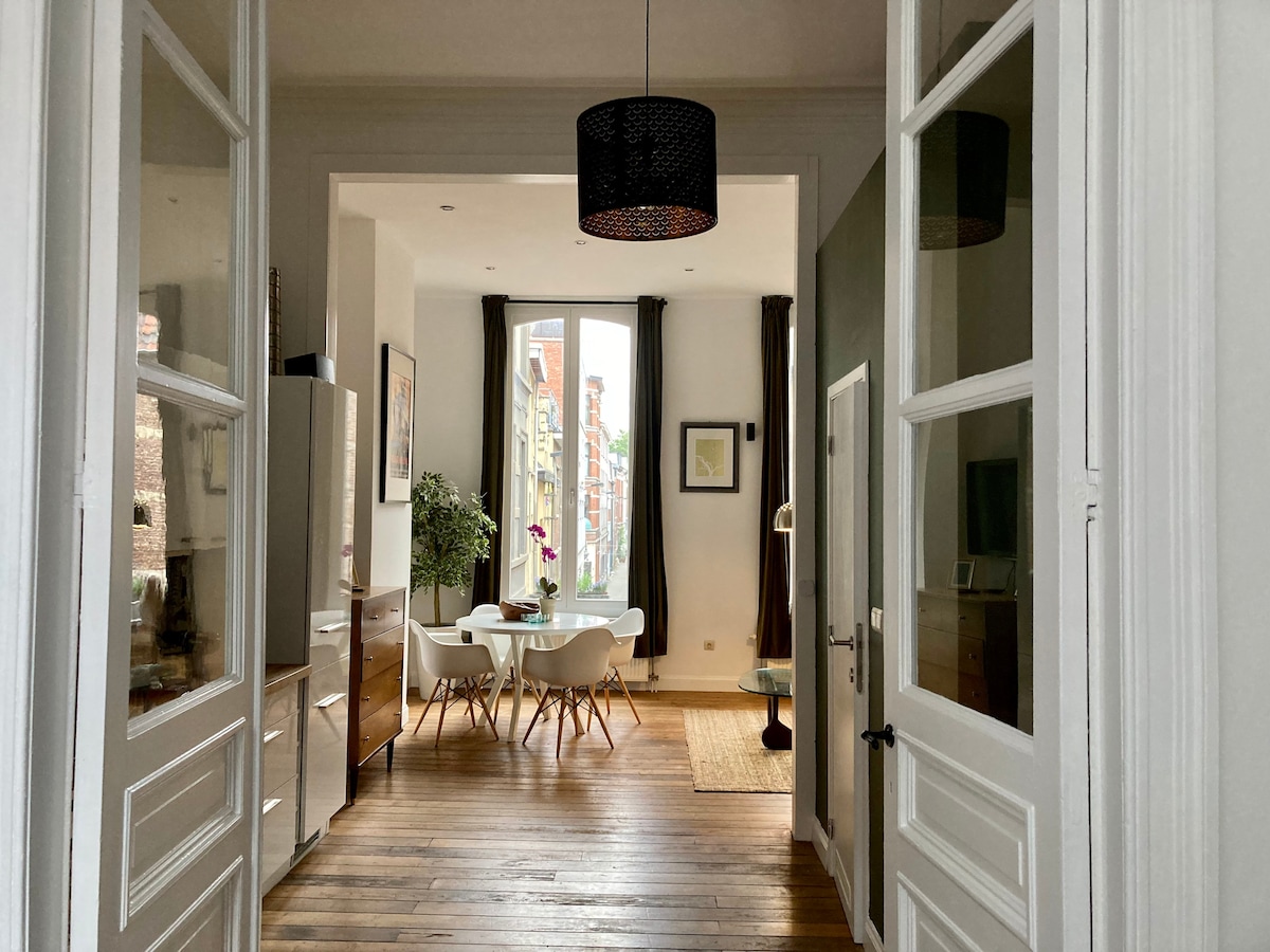 An inviting entrance displays a view of a light-filled living space. Soft green walls complement wood flooring and large windows. A dining area with a circular table and chairs is visible, along with modern furnishings and a potted plant that adds a touch of greenery.