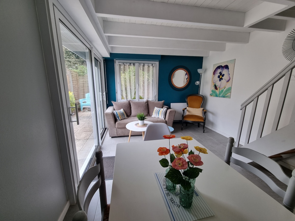 A bright living area features a comfortable sofa and an accent chair, complemented by a round coffee table. Large glass doors open to the garden, allowing natural light to fill the space. Colorful artwork and decorative flowers add a touch of vibrancy.