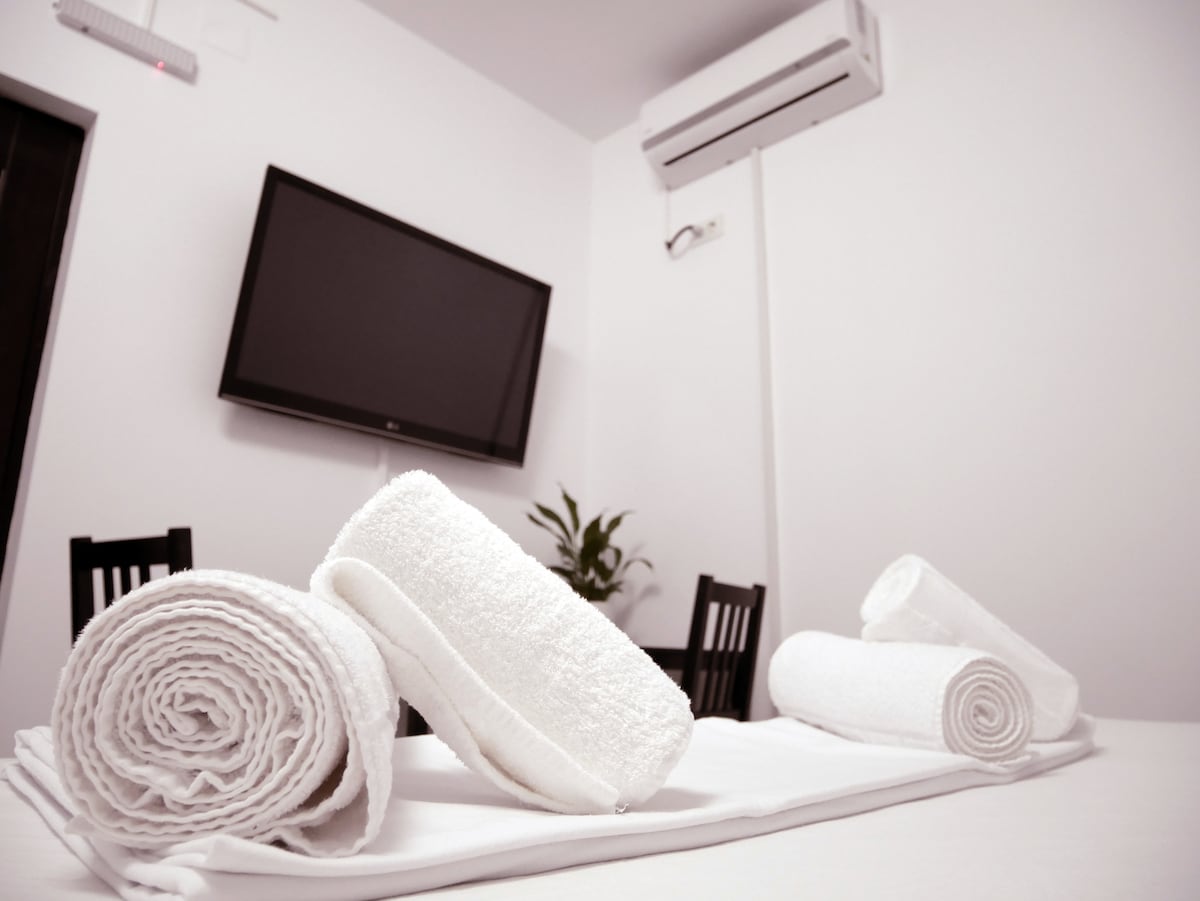 A clean, minimalist space is showcased, featuring neatly rolled white towels on a white surface. A dark wood table with chairs is positioned in the background, while a television is mounted on the wall above. A small green plant adds a touch of color to the area.