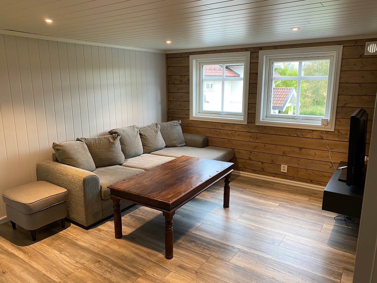 A comfortable living area features a sectional sofa in soft grey positioned near a wooden coffee table. Natural light enters through two windows, with warm wood paneling and light-colored walls creating a cozy ambiance. A flat-screen TV is mounted on a wall opposite the seating.