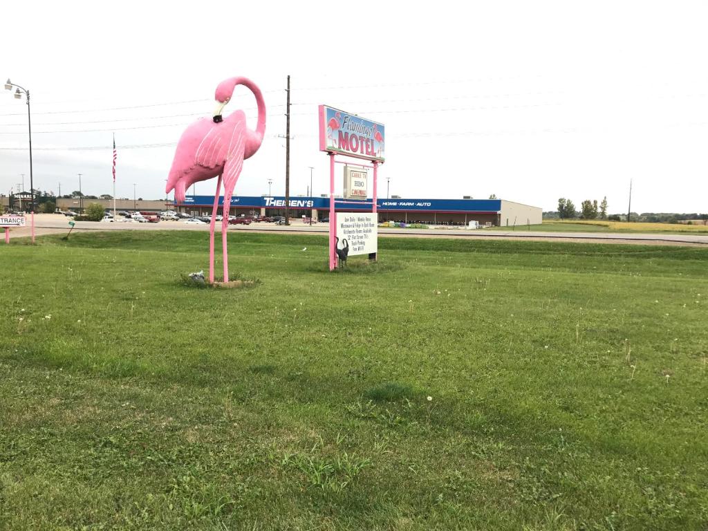 A large pink flamingo sculpture stands prominently in front of the motel, alongside a colorful sign displaying the motel's name. The well-manicured grassy area is visible, creating a welcoming appearance. In the background, several commercial buildings are seen.