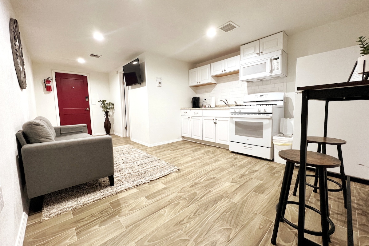 A well-organized kitchen area features white cabinetry, a stove, oven, microwave, and a refrigerator. A cozy seating area is present with a gray couch and a small dining table with wooden stools. Warm wood-like flooring complements the overall neutral decor.