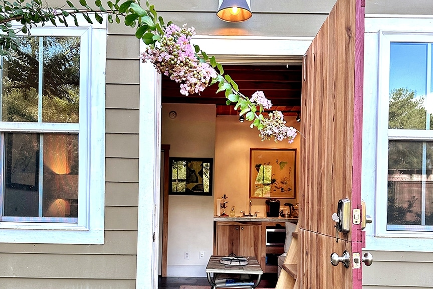 An inviting entrance reveals a glimpse of the interior space. The door swings open to showcase light wood flooring and a view of the kitchen area, framed by two windows. A branch with pink flowers adds a touch of nature to the scene.