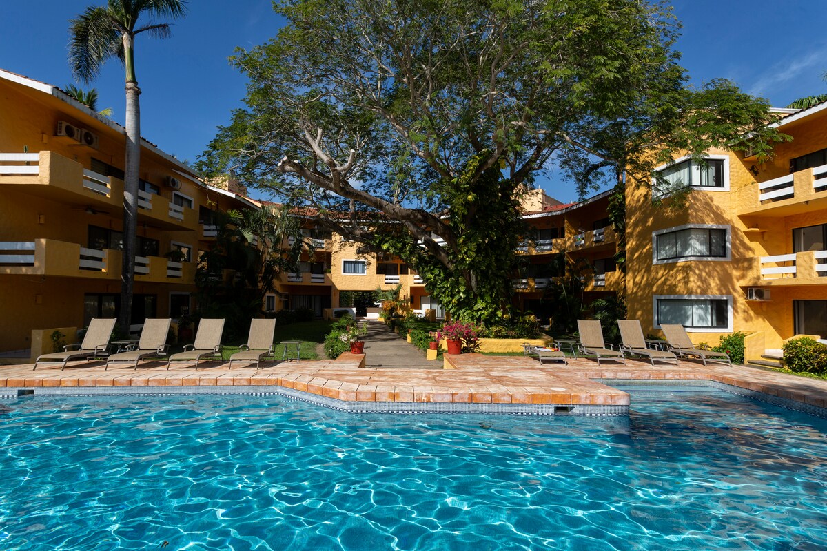 A swimming pool with crystal clear water is surrounded by loungers and lush greenery. The two-story building in the background features multiple windows, and a large tree provides shade, creating a serene outdoor space for relaxation.