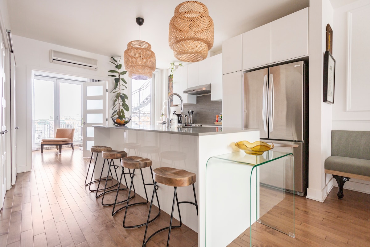 A modern kitchen space features a central island with wooden stools and pendant lighting. High-end appliances are present, complemented by a large refrigerator. Bright natural light fills the area through floor-to-ceiling windows, with a cozy lounge chair positioned nearby.