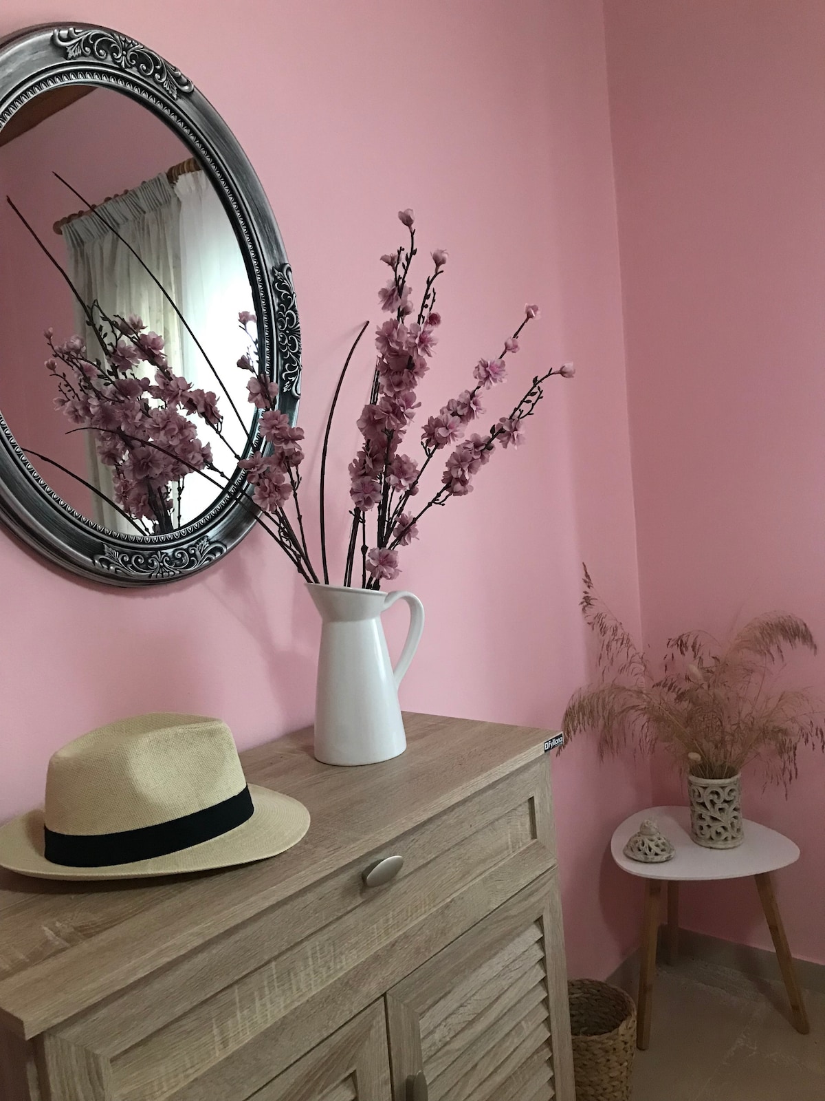 A soft pink wall serves as a backdrop for a wooden cabinet topped with a white pitcher holding pink flowers. A straw hat rests on the cabinet. To the right, a small table displays dried grass and a decorative vase, adding a touch of nature to the space.