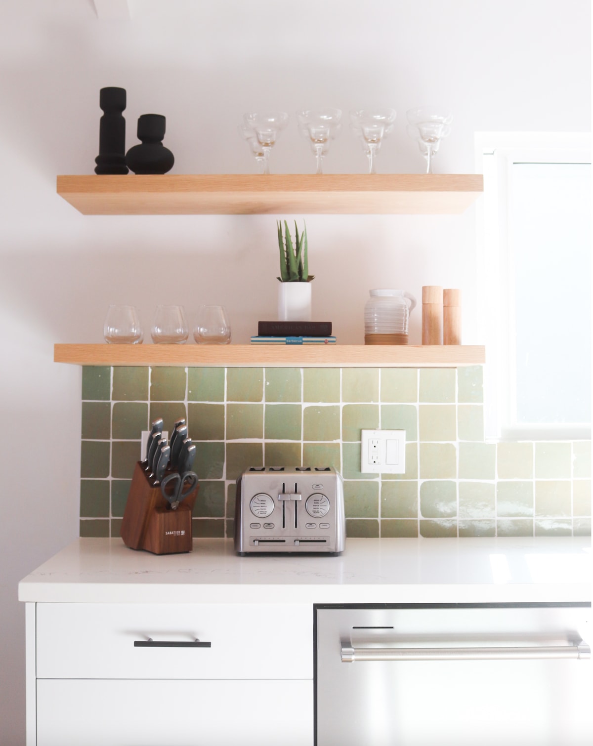 A modern kitchen area features light green tiled backsplash above a white countertop. Two wooden shelves hold glassware and a small potted plant. A stylish toaster sits among kitchen utensils, organized in a wooden block. Natural light brightens the space through a window.