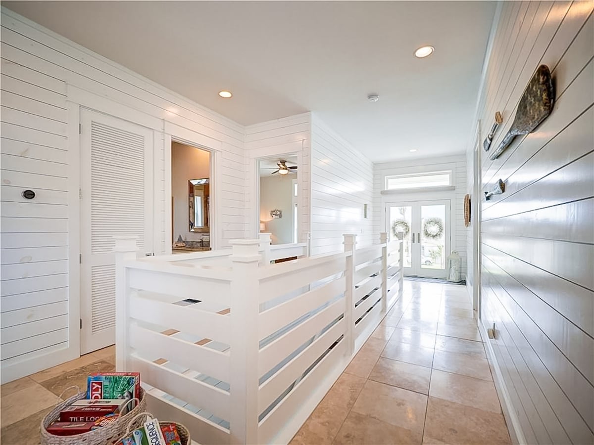 A bright, airy entryway features white shiplap walls and tiled flooring, creating a spacious ambiance. Light streams in through the large front doors, which are flanked by decorative wreaths. A basket containing magazines is positioned on the floor, inviting relaxation.