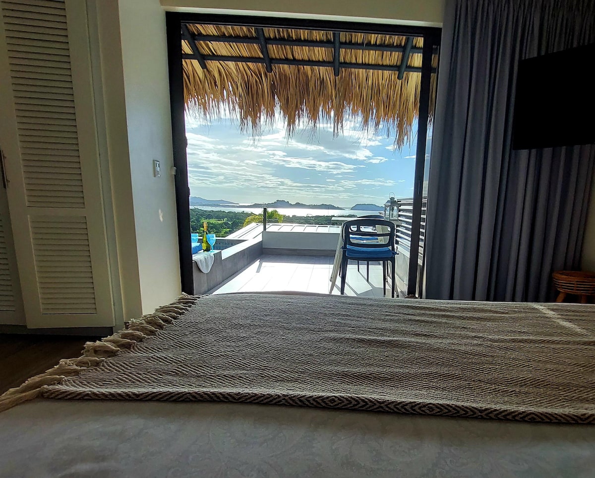 A welcoming view from the spacious bedroom reveals a private terrace, framed by a thatched-roof overhang. Natural light floods the room, highlighting the inviting decor and scenic backdrop of mountains and the ocean beyond.
