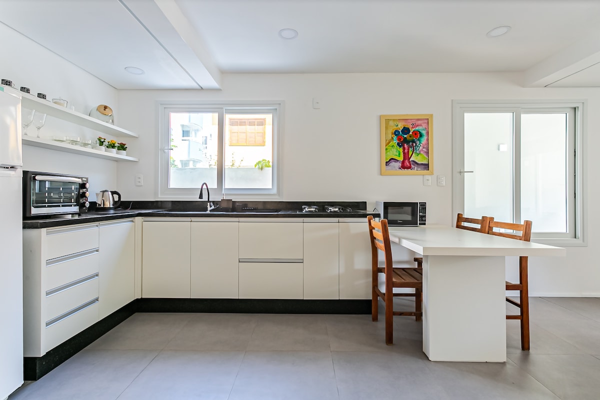 A modern kitchen features sleek white cabinetry and a black countertop. A window allows natural light to brighten the space, alongside a colorful artwork adding a pop of color. A small table with two wooden chairs complements the efficient design.