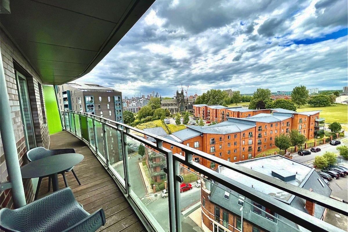 A spacious balcony offers a panoramic view of the surrounding area, including various buildings and green spaces. The railing is made of glass, allowing unobstructed sightlines. Two outdoor chairs are positioned beside a small table, creating a welcoming spot for relaxation.