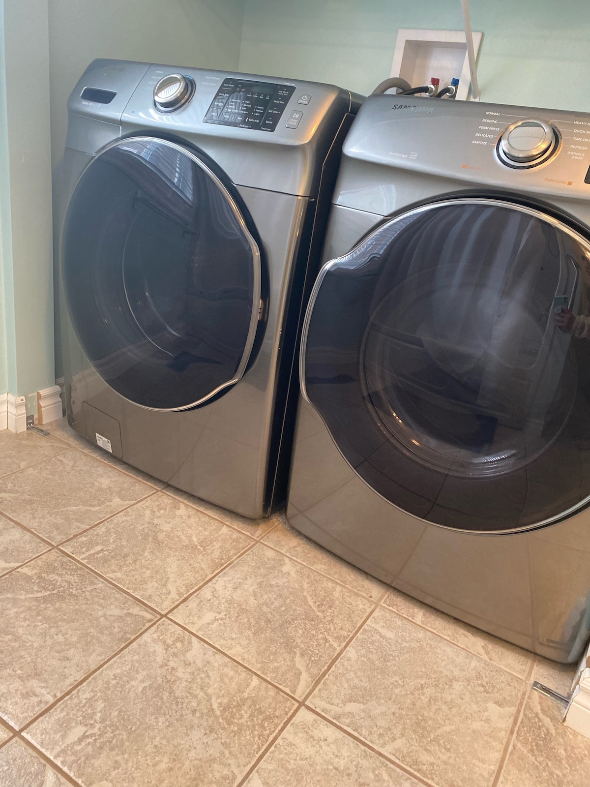 A set of modern washing and drying machines is situated against a wall in a light-colored room. The machines feature a sleek design with large, transparent doors, showcasing the interior. Gray tiled flooring complements the appliances.