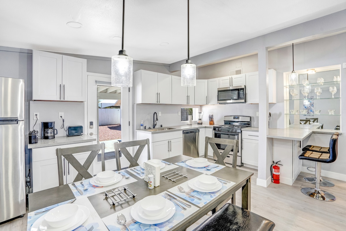 A modern kitchen and dining area are showcased, featuring sleek white cabinetry and stainless steel appliances. A dining table with a blue floral design is set for six, while pendant lights illuminate the space. A bar area is visible with high-backed seating and a clear countertop.