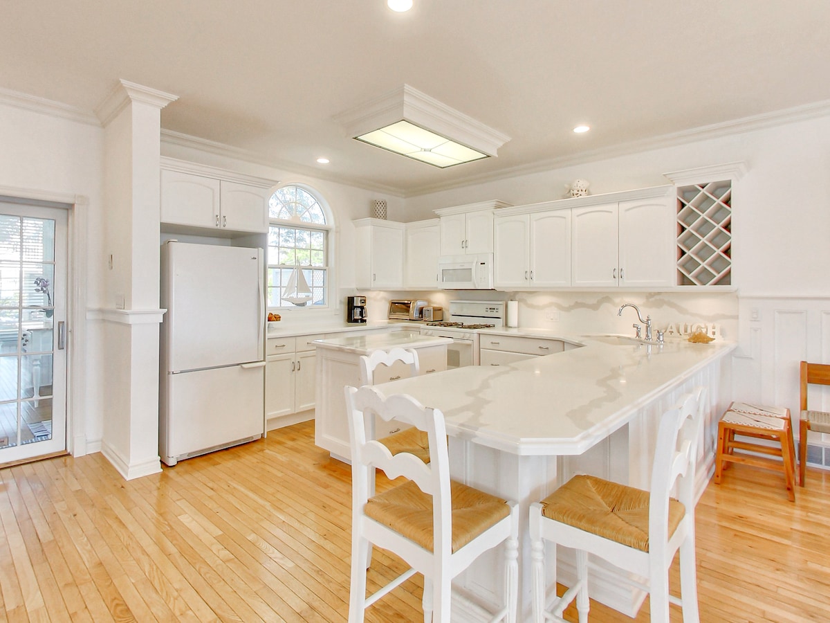 A modern kitchen is presented with bright white cabinetry and marble countertops. An island with high stools provides ample space for culinary activities. Large windows allow natural light to fill the room, highlighting the open layout and inviting atmosphere.