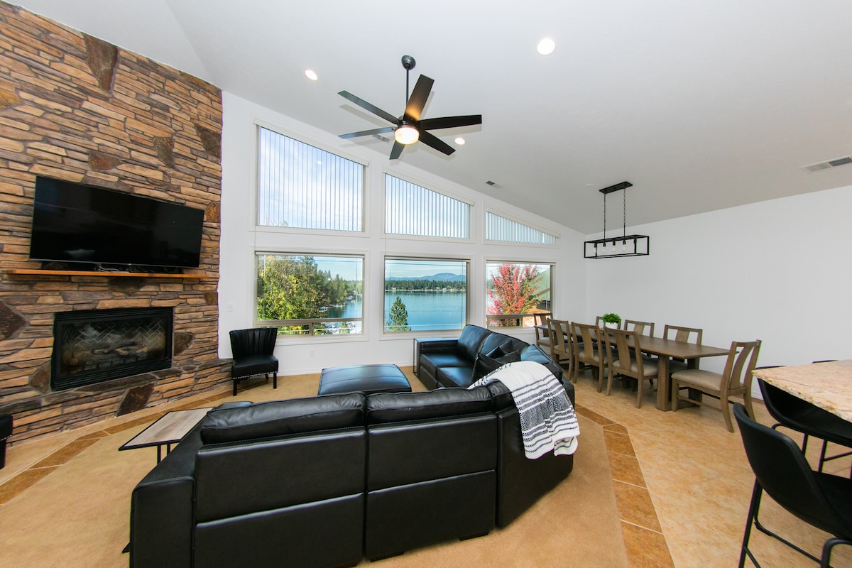 Spacious living area features a black leather sectional and a stone fireplace. Large windows provide views of the lake and surrounding landscape. Natural light enhances the room, which overlooks a dining area with a wooden table and chairs, all set against a neutral-toned wall.