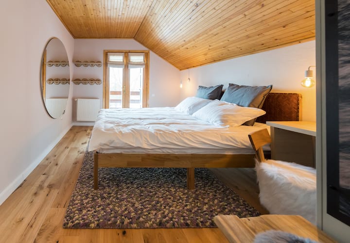 The first bedroom that offers a unique living experience with a queen size bed adorned with white cotton linens, soft pillows for a perfect sleep, modern rug, round mirror, heating radiator, desk with chair for a remote setting and balcony access