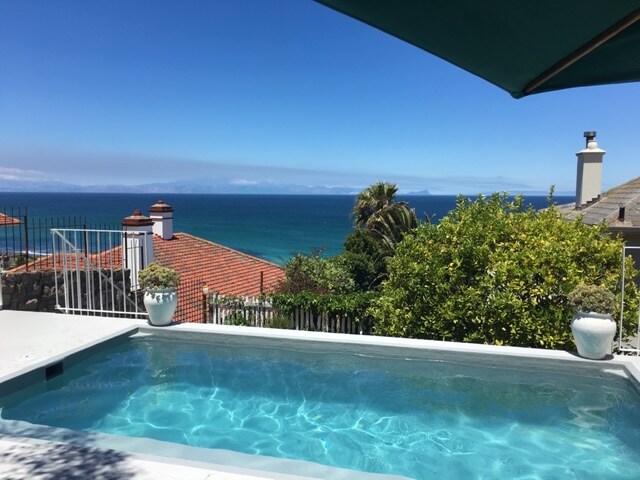 A sparkling swimming pool reflects the clear blue sky and ocean. Lush greenery and well-maintained hedges surround the pool area, providing a sense of privacy. In the distance, the calm sea leads towards the horizon, with rooftops enhancing the seaside view.
