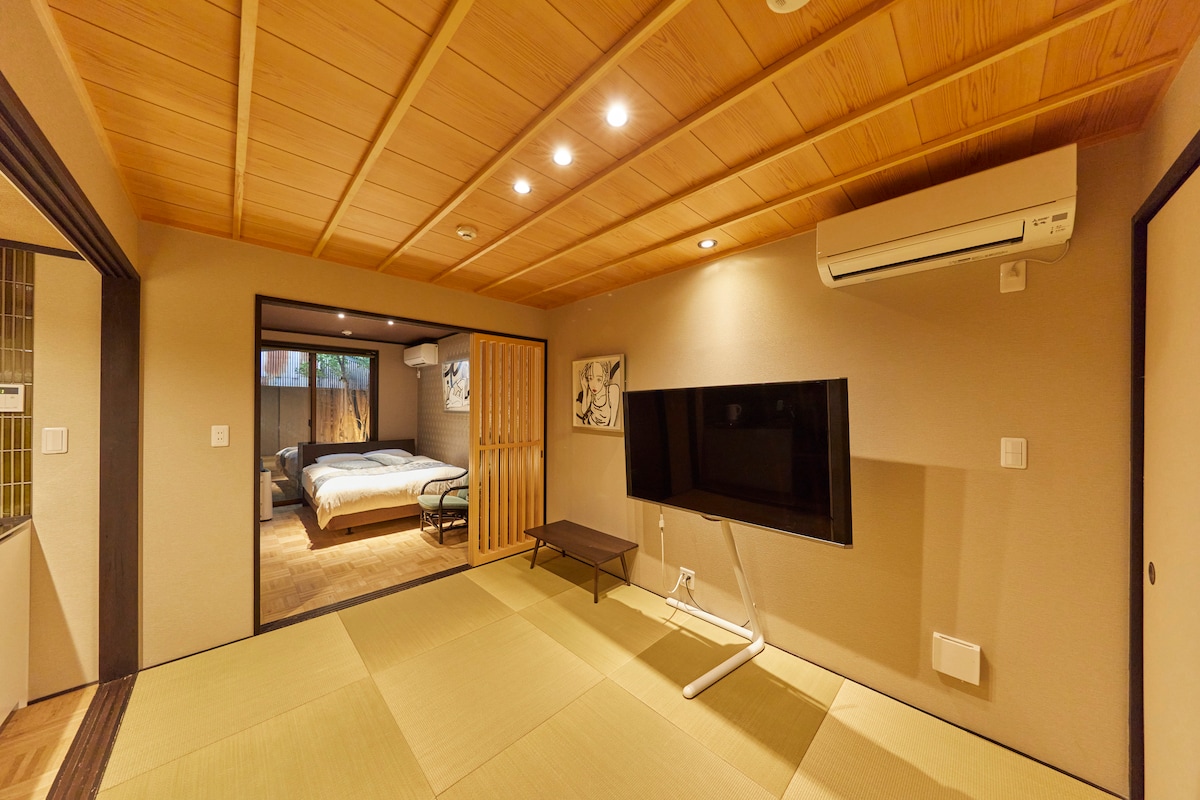 A spacious living area features tatami flooring and wooden ceiling panels. A flat-screen TV is mounted on the wall, while light streams in from the adjoining bedroom. A partition creates a sense of separation, enhancing the room's openness.