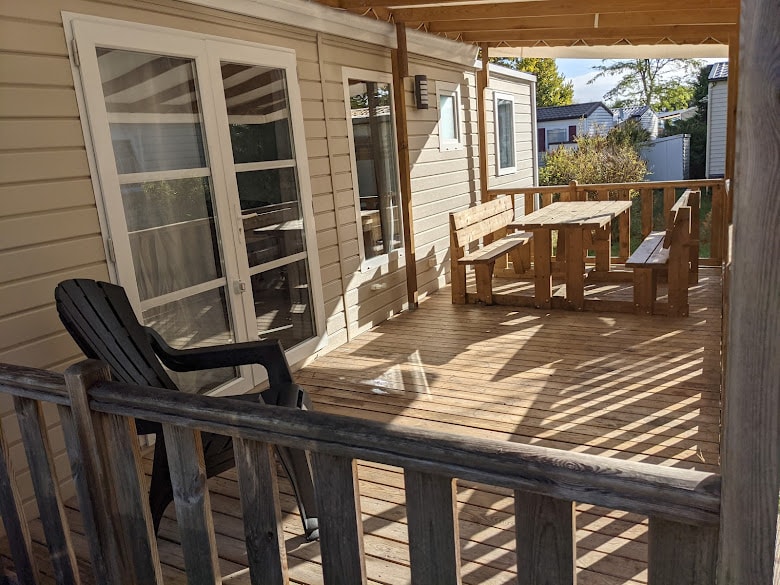 A wooden deck is featured outside the mobile home, showcasing a set of rustic tables and benches arranged for dining. An Adirondack chair is positioned nearby, providing a relaxing spot. Sunlight filters through the overhead beams, casting soft patterns on the floor.