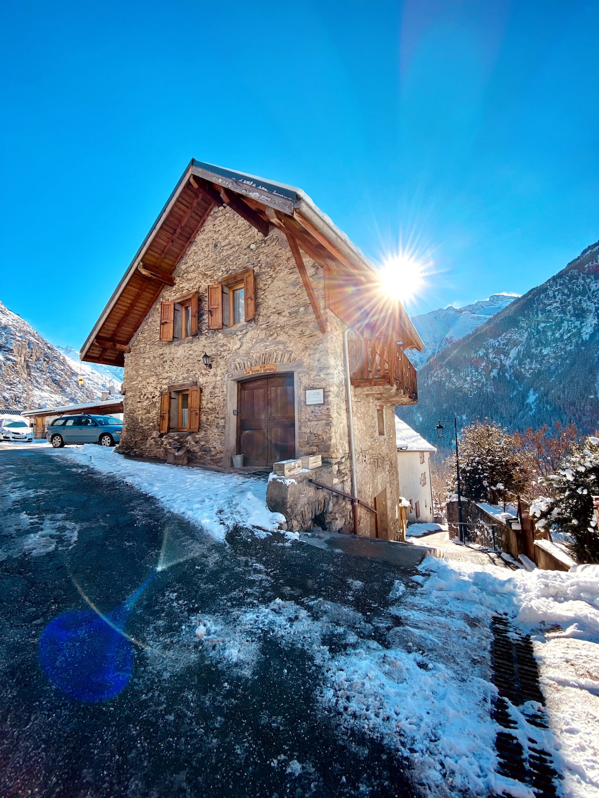 A charming stone chalet is seen amidst a snowy landscape, surrounded by mountains. Sunlight glimmers off the roof, highlighting the wooden shutters and door. The driveway curves gently in front of the chalet, with a vehicle parked nearby, providing an inviting entrance.