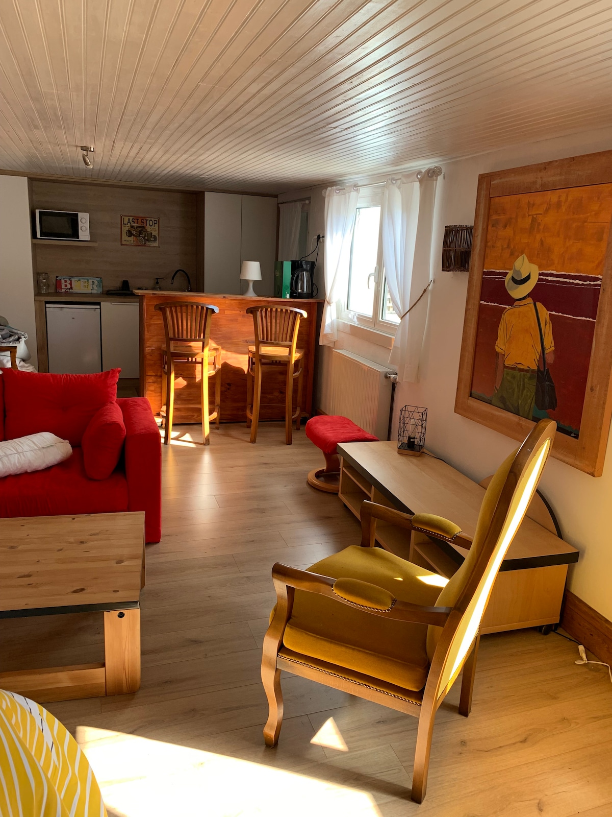 The studio features a cozy living area with a bright red couch and a wooden coffee table. A small kitchenette with bar stools is visible in the background. Natural light floods through the window, highlighting a wooden desk and an armchair near a colorful wall artwork.