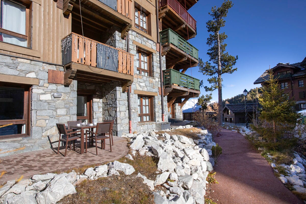 The exterior of the apartment building is showcased, featuring a mix of stone and wood materials. A patio area with outdoor seating is visible, surrounded by landscaped rocky paths and greenery. Balconies with wooden railings are positioned above, offering potential elevated views.