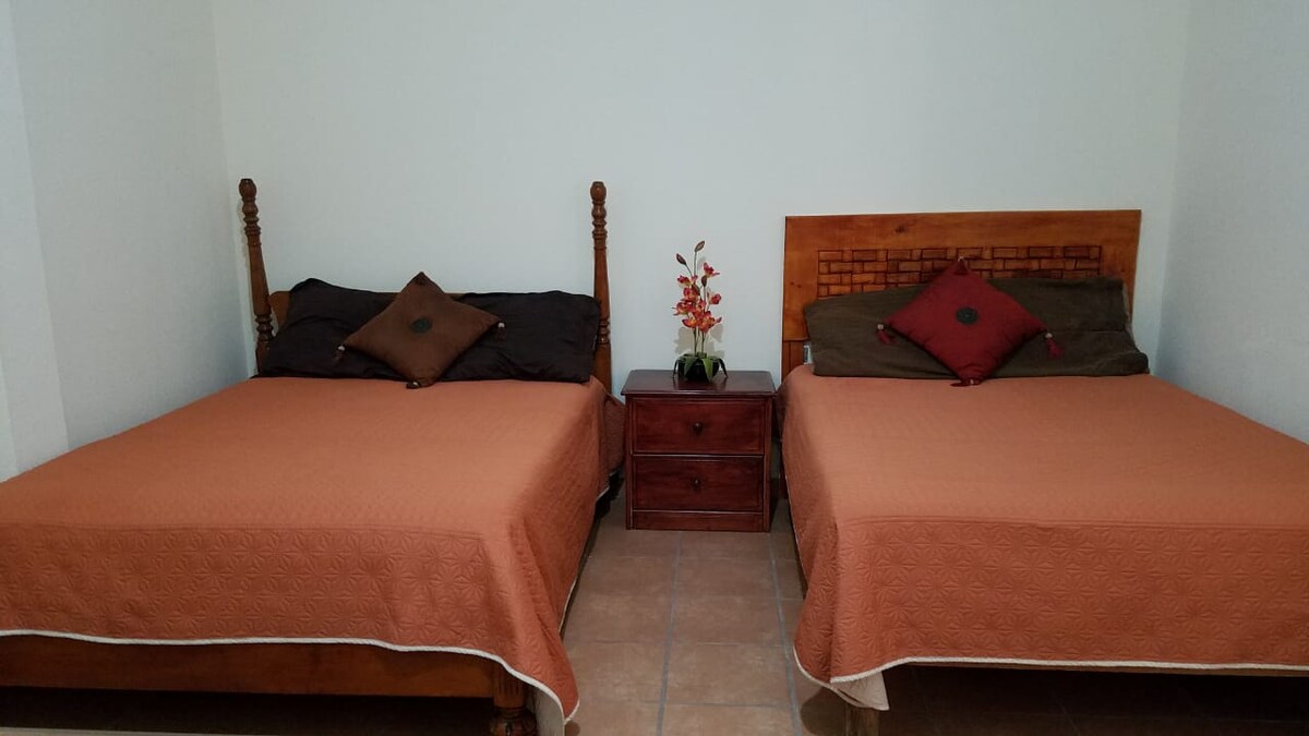 Two spacious beds are positioned in a well-lit room, each adorned with decorative pillows. A nightstand with a small arrangement of flowers is placed between the beds, adding a touch of color to the neutral space. The floor features tiled flooring.