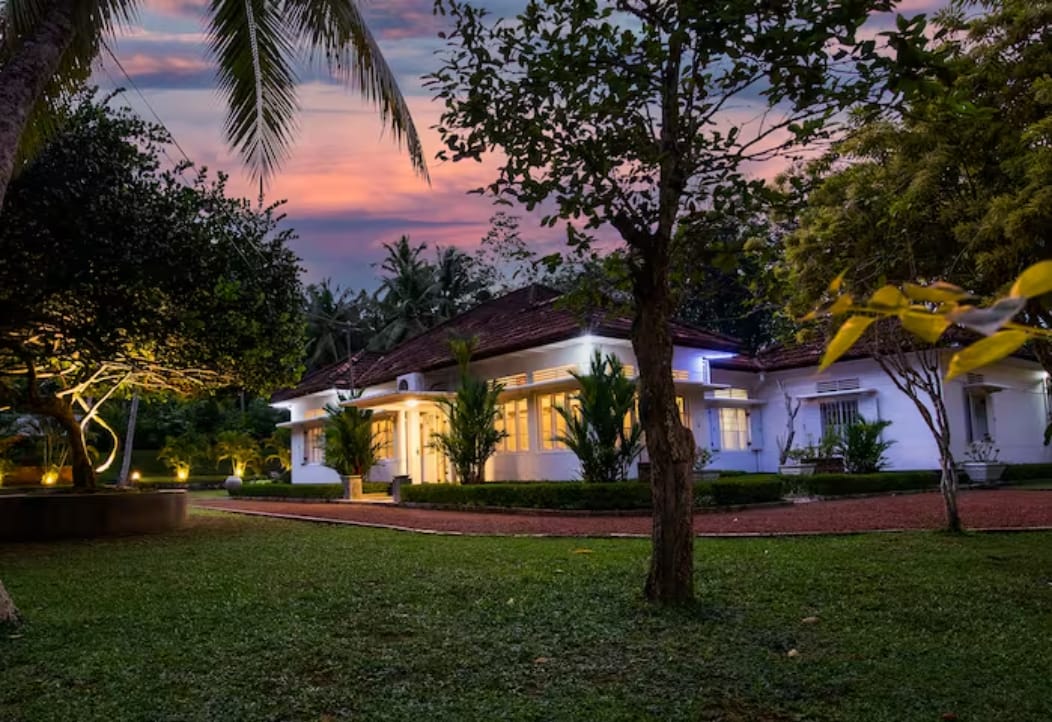 A charming white building is set against a backdrop of palm trees and evening sky hues. Soft outdoor lights illuminate the paths and gardens, while neatly trimmed hedges and manicured lawns frame the peaceful entrance.